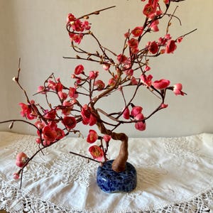 May include: A decorative artificial bonsai tree with red blossoms and brown branches. The tree is set in a blue ceramic pot with a speckled design. The bonsai is placed on a white lace tablecloth.