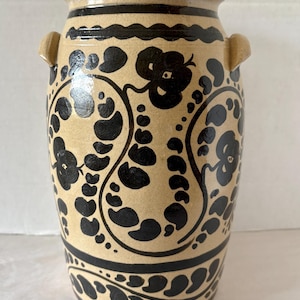 May include: A beige ceramic vase with black floral patterns and two small handles. The vase has a wide opening and a decorative black band around the top and bottom. The floral design includes paisley-like shapes and flower-like motifs.