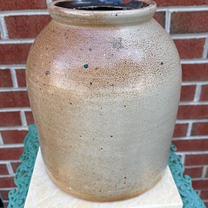 May include: A cylindrical stoneware crock featuring a two-tone glaze with a light beige lower section and a speckled tan upper section. The letter H is stamped into the upper body. The vessel has a thick, rounded rim with a small chip on the edge.