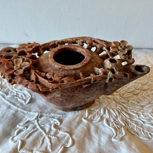 Könnte beinhalten: Eine handgeschnitzte Öllampe aus rotem und braunem Stein mit aufwendigen floralen Designs. Die Lampe hat einen Ausguss und eine runde Öffnung für einen Docht.