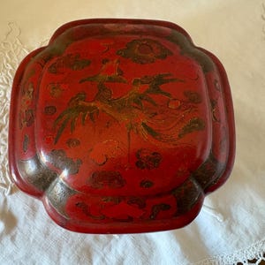 May include: A red, antique decorative tray featuring a bird and cloud design. The tray has a unique shape with rounded edges and a slightly raised rim. The bird is depicted in gold and brown tones, contrasting the red background.