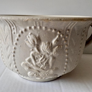 May include: A beige ceramic chamber pot featuring a raised relief design of flowers on the front. The vessel has a wide, flared rim, a single side handle, and vertical textured panels that frame the central floral motif with beaded borders.