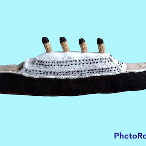 May include: A handcrafted toy ship in black, white, and beige. The ship has four smokestacks, a white upper deck with black dot details, and the word "TITANIC" on the side. The toy is set against a light blue background.
