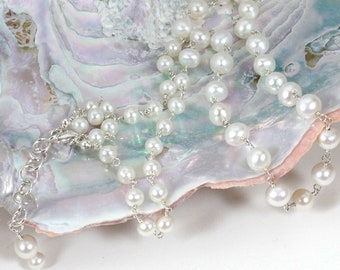 May include: A delicate pearl necklace with a silver chain and clasp. The necklace features multiple strands of white pearls of varying sizes, creating a layered effect. The jewelry is displayed on a pearlescent, iridescent surface.