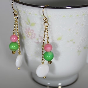 Op de afbeelding: Goudkleurige oorhangers met roze, groene en witte kralen. De oorbellen hebben een kettingontwerp en een haaksluiting. De kralen zijn rond en ovaal, met een glanzende afwerking. De oorbellen worden gepresenteerd voor een witte theekop.