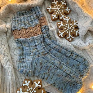 Peut inclure: Une paire de chaussettes tricotées bleues et marron avec une bande décorative marron, posée sur une couverture crème. Trois biscuits en pain d'épices en forme de flocon de neige avec du glaçage blanc sont également présents.