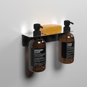 Op de afbeelding: Een zwarte metalen wanddispenser met twee amberkleurige glazen flessen met pompen. De flessen hebben etiketten met de tekst "Shampoo/Scianpo" en "Conditioner/Balsamo". Een gele zeepstaaf zit in de dispenser.