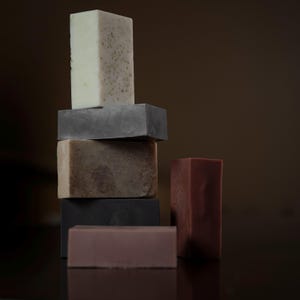 May include: A stack of handmade soap bars in various colors, including white, gray, brown, and reddish-brown. The soaps are rectangular and have a natural, textured appearance, suggesting a focus on organic ingredients and artisanal craftsmanship.