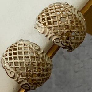 May include: Two ornate, spherical earrings. Each earring features a lattice design with a cream-colored finish and gold accents. The earrings are displayed on a gold-colored rod against a neutral background.