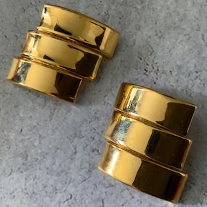 May include: Two gold-colored, stacked cuff bracelets. Each bracelet has three curved, rectangular bands. The bracelets are on a light gray surface. The gold color is shiny and reflective.