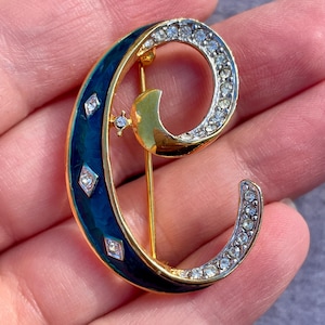 May include: A gold-coloured brooch in the shape of the letter "C", with a crescent moon and star design. The brooch features a dark blue enamel section with diamond-shaped accents and a section encrusted with small, clear crystals. A gold pin is attached to the back.
