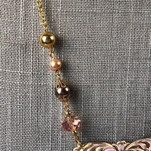 May include: A gold-toned necklace with a delicate chain and decorative beads. The necklace features a large gold bead, a smaller peach-colored bead, a dark brown bead with filigree detailing, and a pink crystal. The necklace is set against a gray textured background.