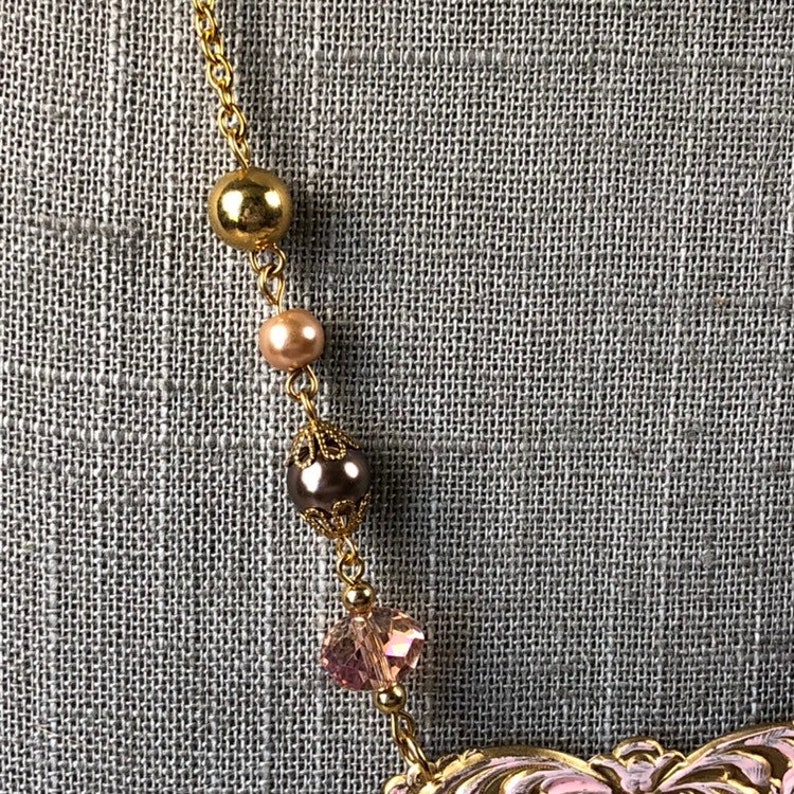 May include: A gold-toned necklace with a delicate chain and decorative beads. The necklace features a large gold bead, a smaller peach-colored bead, a dark brown bead with filigree detailing, and a pink crystal. The necklace is set against a gray textured background.