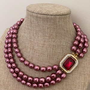 May include: A three-strand necklace featuring lustrous mauve-colored pearls. The strands are connected by a rectangular gold-toned clasp set with a large, deep red rectangular gemstone surrounded by a border of small, clear white crystals.