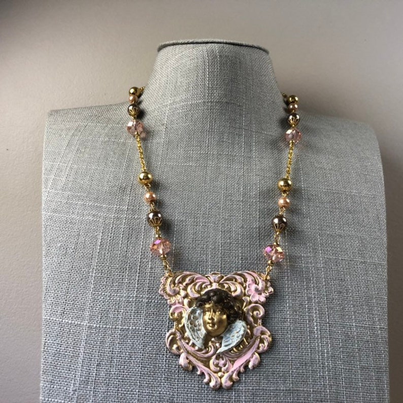 May include: A gold-toned necklace featuring a pendant with a cherub face. The pendant is framed in pink and gold filigree. The necklace has gold beads and pink crystal accents. The necklace is displayed on a gray mannequin.