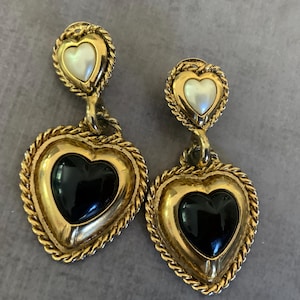 May include: A pair of gold-toned heart-shaped earrings. Each earring features a small, white heart at the top and a larger, black heart at the bottom. The edges are adorned with a rope-like design.