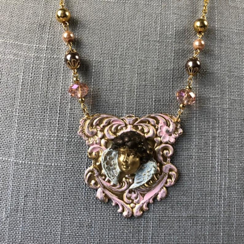 May include: A gold-toned necklace featuring a pendant with a cherub face. The pendant is framed in pink and gold filigree. The necklace chain has gold beads, pink pearls, and faceted crystals.