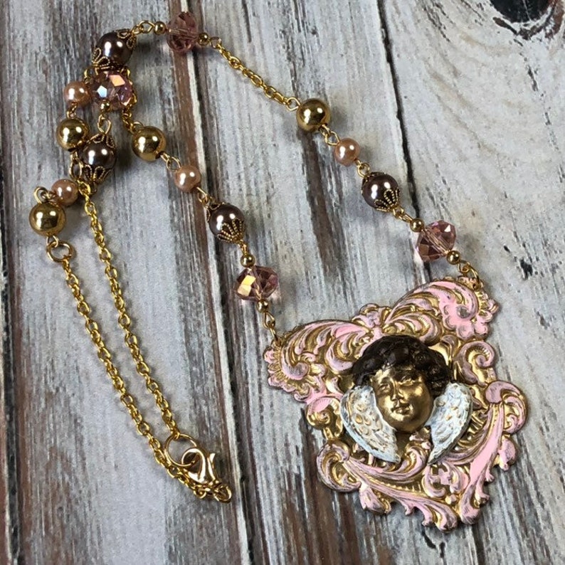 May include: A gold-toned necklace featuring a pendant with a cherub face and wings, set within a pink and gold ornate frame. The necklace includes gold chain, pink and brown beads, and crystal accents, creating a vintage aesthetic.