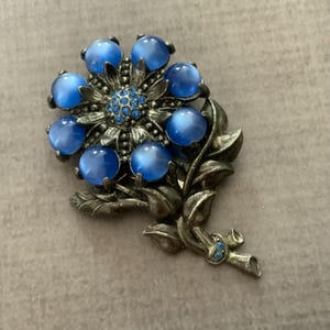 May include: A floral brooch featuring seven round, translucent blue cabochon petals surrounding a center of small, sparkling blue rhinestones. The flower is attached to a textured, antiqued silver-tone metal stem with two leaves.