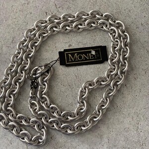 May include: A silver-toned chain necklace featuring thick, textured oval links. A black rectangular tag with the gold-colored text MONET is attached to the chain by a thin black cord. The necklace has a lobster claw clasp closure.