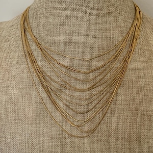 May include: A multi-strand gold-toned necklace featuring numerous delicate, thin chains that drape in a layered, cascading fashion. The chains are uniform in color and texture, creating a layered effect that rests against the neck area.