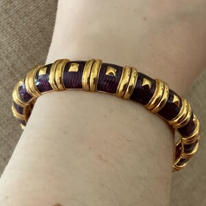 May include: A hinged bangle bracelet featuring alternating segments of polished gold-tone metal and deep purple enamel with a subtle wavy texture. Each purple section is accented with a small, raised gold-tone square stud detail.