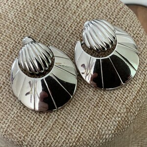 May include: A pair of silver earrings. Each earring has a ribbed, rounded top and a crescent-shaped bottom with radiating lines. The earrings are displayed on a textured, neutral-coloured surface.
