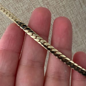 May include: A gold-colored herringbone chain necklace. The chain is flat and has a woven pattern. The necklace is held in a person's hand, with the chain extending diagonally across the frame. The background is a neutral beige color.