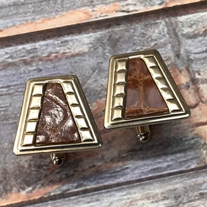 Puede incluir: Un par de gemelos de oro con una incrustaci&oacute;n de cuero marr&oacute;n. Los gemelos tienen forma de trapecio con un borde dorado.