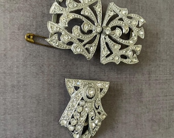 May include: A silver-toned metal dress clip set featuring two pieces with intricate openwork filigree designs. Both pieces are encrusted with clear, sparkling rhinestones. One piece is a larger, wider clip, and the other is a smaller, tapered clip.