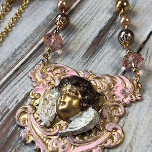 May include: A gold-toned necklace featuring a pendant with a cherub face and wings. The pendant is set against a pink and gold filigree design. The necklace includes pink crystal beads and gold-colored accents.