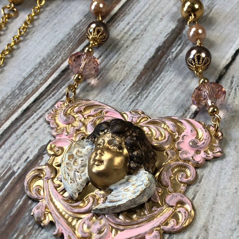 May include: A gold-toned necklace featuring a pendant with a cherub face and wings. The pendant is set against a pink and gold filigree design. The necklace includes pink crystal beads and gold-colored accents.