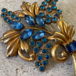 May include: Vintage gold-toned brooch featuring a floral design with blue crystal accents. The brooch has a cluster of round and oval blue crystals, resembling grapes, and is set against gold-colored leaves.