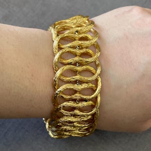 May include: A wide gold-tone bracelet featuring a series of interconnected, textured oval links. The metal surface has a brushed, organic finish, creating a repeating pattern of horizontal loops that wrap around the wrist in a structured, openwork design.