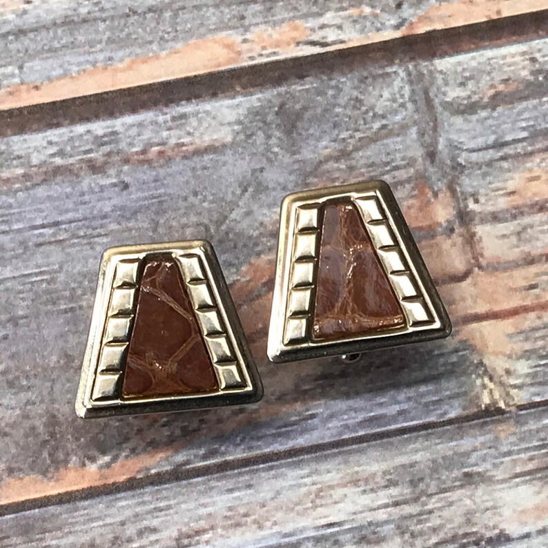 Puede incluir: Un par de pendientes de tono dorado con una incrustaci&oacute;n de imitaci&oacute;n de cuero marr&oacute;n. Los pendientes tienen forma de trapecio con un borde dorado y un patr&oacute;n texturizado.