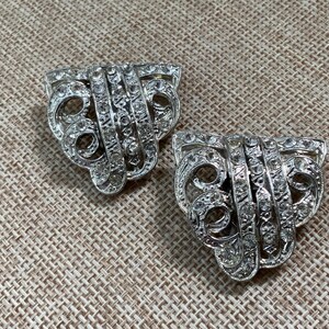 May include: Two silver-toned Art Deco style dress clips. Each clip has a fan-shaped design with a central vertical band and curved, openwork sides, all encrusted with clear rhinestones. The clips are displayed on a textured, neutral background.