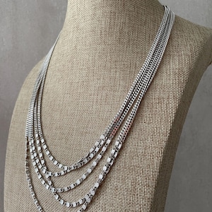 May include: A multi-strand silver-toned necklace featuring four distinct layers of chain. The design includes two upper strands of fine, tightly linked metal chains and two lower strands of textured, oval-shaped links that create a layered appearance.
