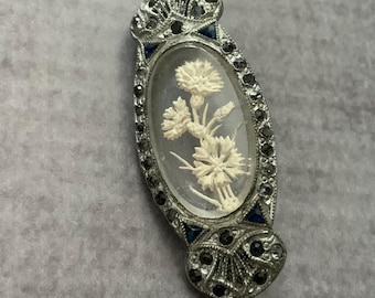 May include: An oval brooch featuring a central clear glass cabochon with an off-white carved floral motif. The frame is crafted from silver-toned metal, accented with small dark grey stones and four deep blue triangular stones at the edges.