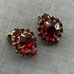 May include: A pair of vintage clip-on earrings. Each earring features a large, round, faceted, deep red stone surrounded by smaller red stones. The setting is gold-toned metal, and the earrings are displayed on a gray surface.