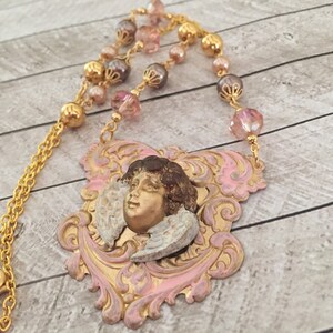 May include: A gold-colored necklace featuring a pendant with a cherub face and wings. The pendant is framed by pink and gold filigree. The necklace chain is gold, with pink, gold, and brown beads.