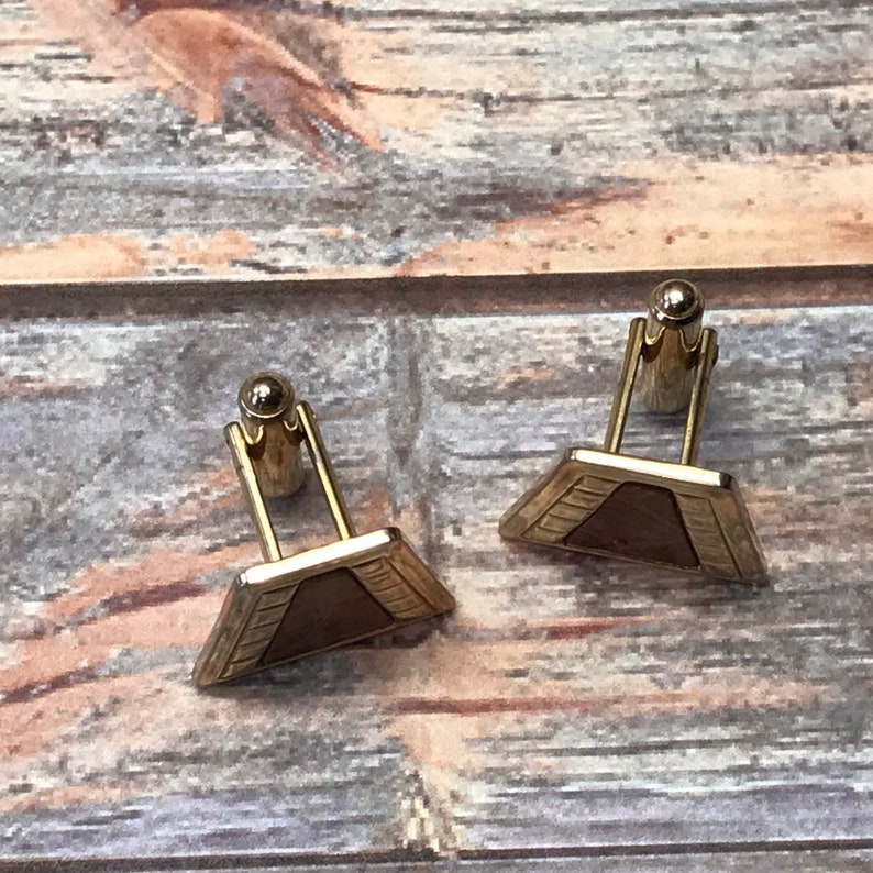 Puede incluir: Un par de gemelos de tono dorado con un dise&ntilde;o triangular. El centro de cada gemelo es de un material marr&oacute;n similar a la madera.