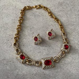 May include: A gold-tone necklace and matching clip-on earring set featuring square-shaped links adorned with clear rhinestones and a central oval-cut red gemstone. The necklace chain consists of interlocking gold-tone metal links.