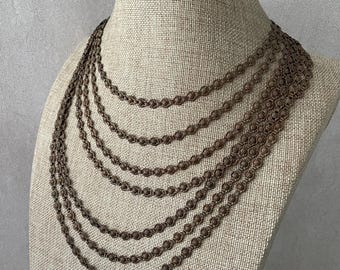 May include: A multi-strand necklace featuring five cascading layers of dark bronze-toned chain. Each chain is composed of small, repeating circular links with a textured, floral-like pattern, creating a uniform and layered appearance.