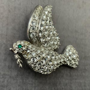 May include: A silver-toned dove brooch encrusted with clear rhinestones. The dove holds a sprig in its beak and has a green gemstone eye. The wings are outstretched, and the brooch is set against a gray background.