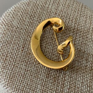 May include: A gold-colored brooch in the shape of the letter "G". The pin is made of a shiny, reflective metal and is set against a neutral-toned, textured background. The brooch has a classic, elegant design.