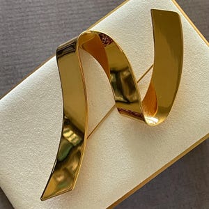 May include: A gold-toned brooch with a ribbon-like design. The brooch has a pin on the back for secure fastening. The ribbon curves and folds, creating a dynamic and elegant look. The brooch is likely made of metal and has a polished finish.