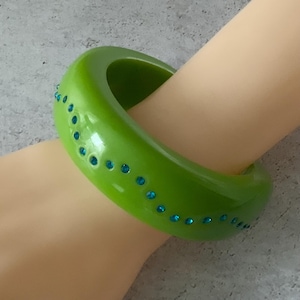 May include: A bright green bangle bracelet with a rounded rectangular shape. The bracelet is adorned with small, round, blue gemstones. The bracelet is worn on a person's arm.