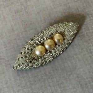 May include: An elongated, leaf-shaped brooch featuring a silver-toned metal frame encrusted with numerous small, clear, sparkling rhinestones. Three smooth, round, cream-colored faux pearls are set in a horizontal row along the center of the piece.