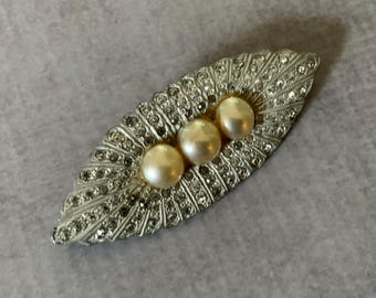 May include: An elongated, leaf-shaped brooch featuring a silver-toned metal frame encrusted with numerous small, clear, sparkling rhinestones. Three smooth, round, cream-colored faux pearls are set in a horizontal row along the center of the piece.