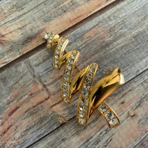 May include: A gold-toned Christmas tree brooch with clear rhinestones. The brooch is shaped like a stylised Christmas tree with a wavy design.